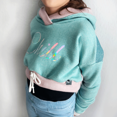 Person in einem Cropped Hoodie von Vorn. Der Pullover hat die Farben Mint, Altrosa und Petrol. Das Muster ist ein leichtes Fischgrätmuster. Hoodie ist vorn mit Schriftzug in geschwungener Schreibschrift "Chill".