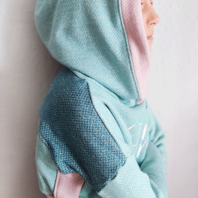 Person in einem Cropped Hoodie mit aufgesetzter Kapuze von der Seite. Der Pullover hat die Farben Mint, Altrosa und Petrol. Das Muster ist ein leichtes Fischgrätmuster.