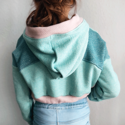 Person in einem Cropped Hoodie von Hinten. Der Pullover hat die Farben Mint, Altrosa und Petrol. Das Muster ist ein leichtes Fischgrätmuster.