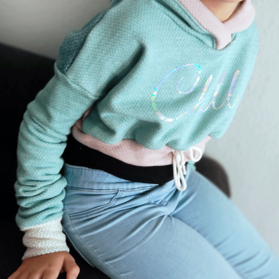 Person auf einem Sofa die einen Hoodie presentiert. Der Pullover hat die Farben Mint, Altrosa und Petrol. Das Muster ist ein leichtes Fischgrätmuster. Der Hoodie hat vorn eine Aufschrift in geschwungener Schreibschrift "Chill".