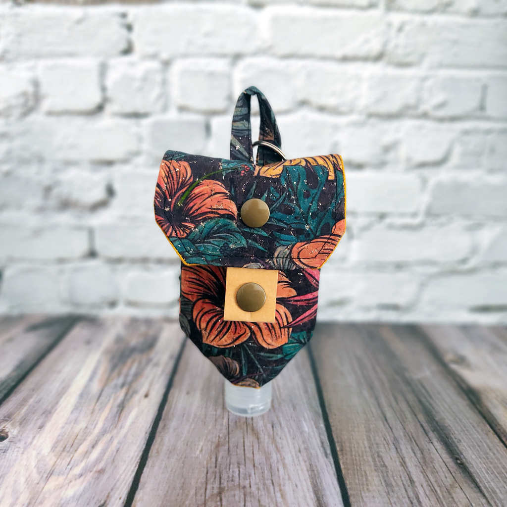 Frontansicht einer Tasche für Desinfektionsmittel, genäht aus Korkstoff mit Blumen und leichtem Glitzer. Die Tasche ist mit einem von zwei Druckknöpfen geschlossen. Bild zeigt die Passvariante für kleine Flaschen.
